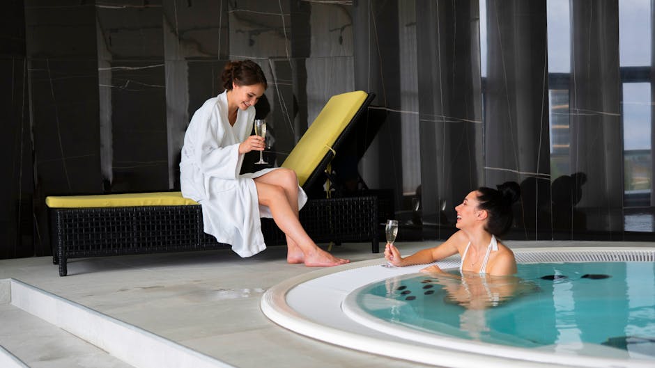 Two women relaxing and bonding with champagne beside an indoor jacuzzi in a spa setting.