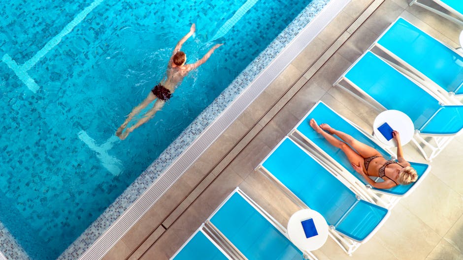 High-angle shot of a swimmer in a pool and a woman sunbathing on a chaise lounge nearby.